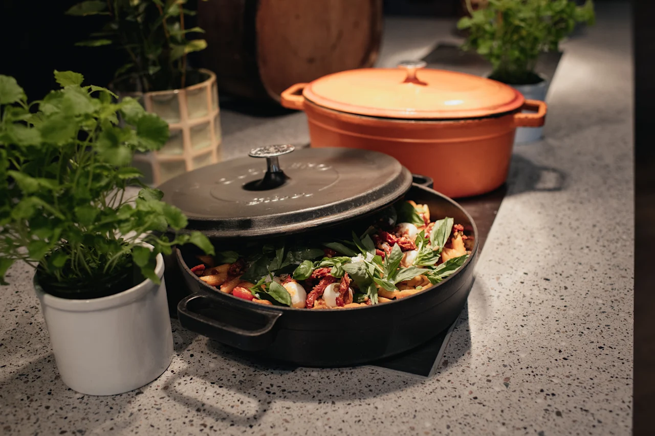 Et deilig måltid tilberedes i en svart gryte, pyntet med fersk basilikum og tomater, ved siden av en oransje gryte og grønne planter.