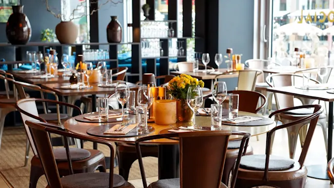 Et dekket bord med blomster og glass, arrangert på trestoler i en koselig, varmt opplyst restaurant med store vinduer og dekorative vaser i bakgrunnen.