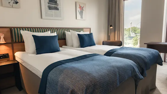 Et lyst hotellrom med to komfortable senger, blå detaljer og utsikt over byen fra vinduet.