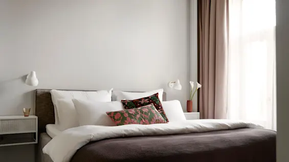 En pent oppreid seng med lagvise puter og et brunt teppe i et mykt opplyst rom, omgitt av veggmonterte lamper og gardinerte vinduer.