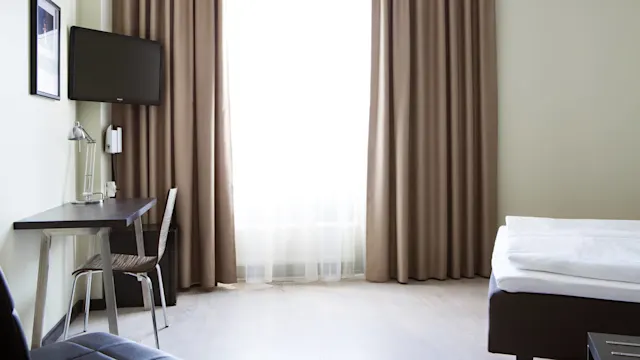 A desk with a lamp and chair sits by a window with beige curtains in a minimalistic room. A bed is partially visible, and a wall-mounted TV is present.