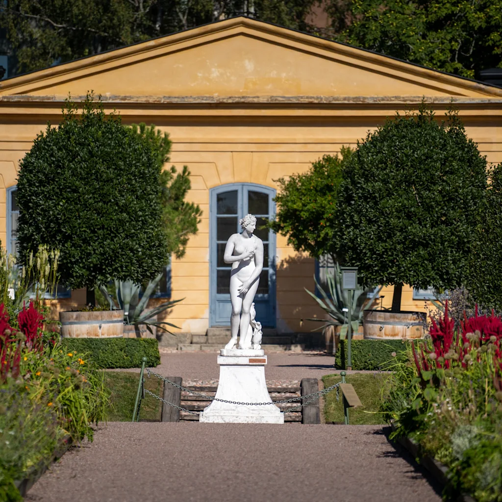 Klassinen patsas seisoo puutarhapolun päässä, johtaen historialliseen keltaiseen rakennukseen Uppsalan Linnéträdgårdenissa.