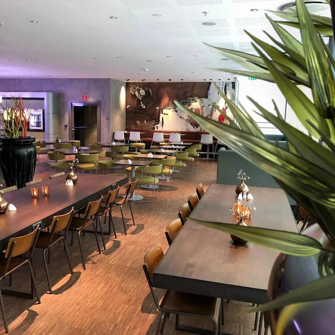 Wooden tables adorned with small decorative vases and candles are surrounded by chairs in a spacious, modern dining area featuring a large potted plant and abstract wall art.