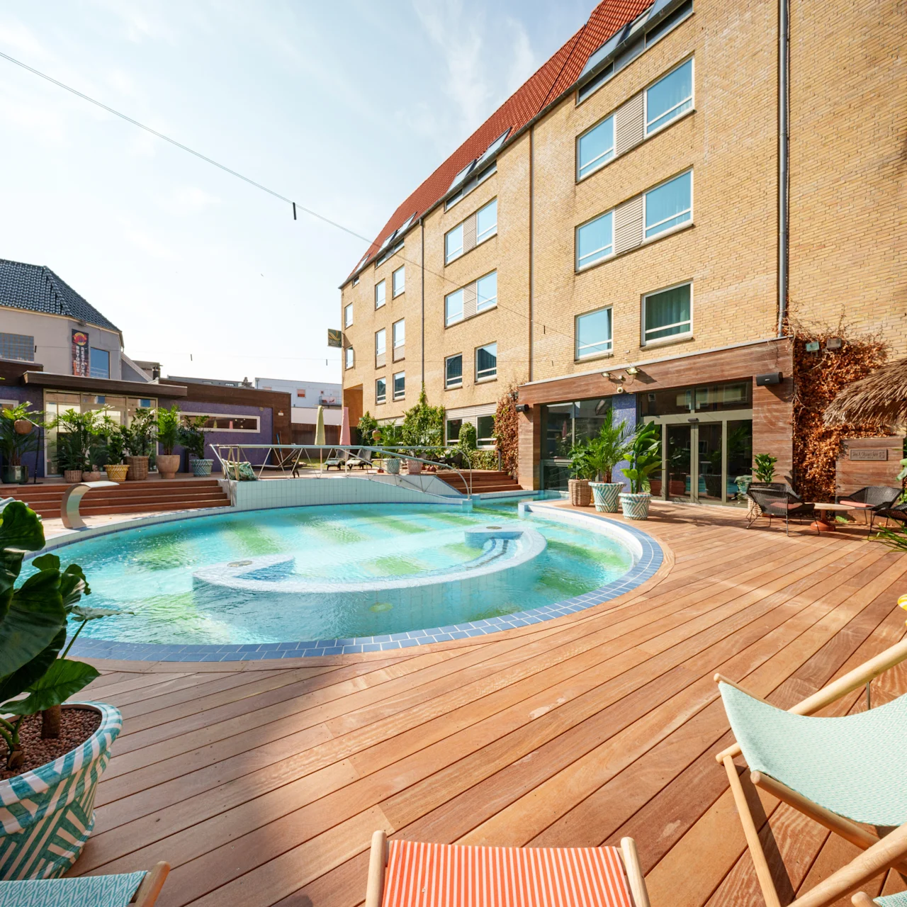 A round swimming pool is surrounded by a wooden deck, lounge chairs, and potted plants, adjacent to a multi-story brick building with large windows.