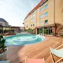 En rund swimmingpool omgives af et trægulv, liggestole og potteplanter, ved siden af en fleretagers murstensbygning med store vinduer.