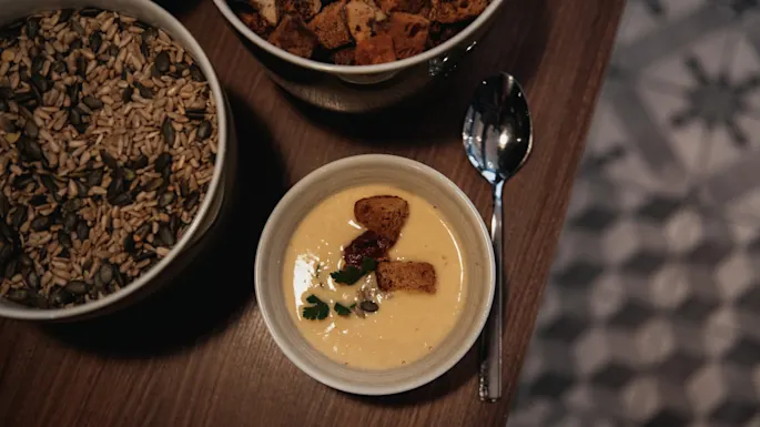 En bolle kremet suppe med krutonger og en skje, akkompagnert av boller med frø og flere krutonger på et trebord.