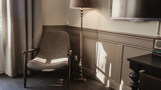 Ett solbelyst hörn av ett hotellrum med en bekväm stol, en golvlampa och en väggmonterad TV, vilket skapar en avkopplande atmosfär.