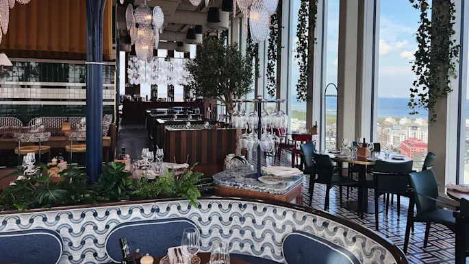 Luxurious restaurant features hanging glass chandeliers and lush greenery. Dining tables are elegantly set near large windows, offering a panoramic city and sea view. Elegant ambiance with detailed decor.