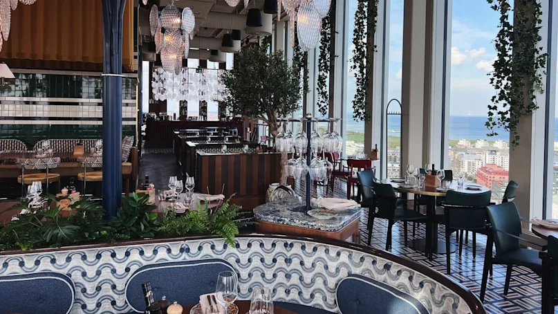 Luxurious restaurant features hanging glass chandeliers and lush greenery. Dining tables are elegantly set near large windows, offering a panoramic city and sea view. Elegant ambiance with detailed decor.