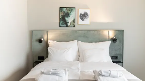 Viihtyisä hotellihuone siististi pedatulla sängyllä, valkoisilla liinavaatteilla ja kahdella taideteoksella sängynpäädyn yläpuolella, valmiina vieraalle.