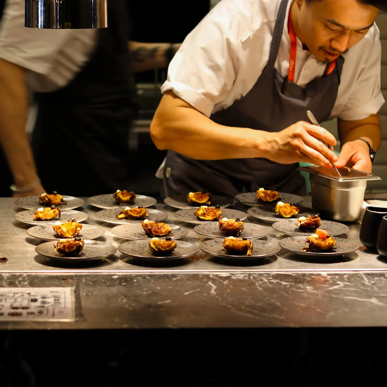 Chef plating dishes meticulously in a dimly lit kitchen, surrounded by plates of food, sauce bottles, and cooking equipment, emphasizing culinary precision and attention to detail.