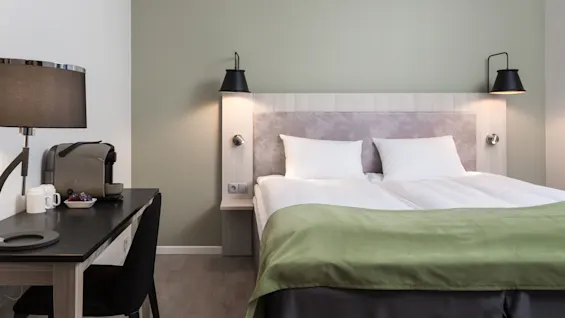 A neatly made bed with white pillows and a green bedspread. A desk with a coffee machine, cups, and a lamp is to the side. The room has a modern, minimalist design.