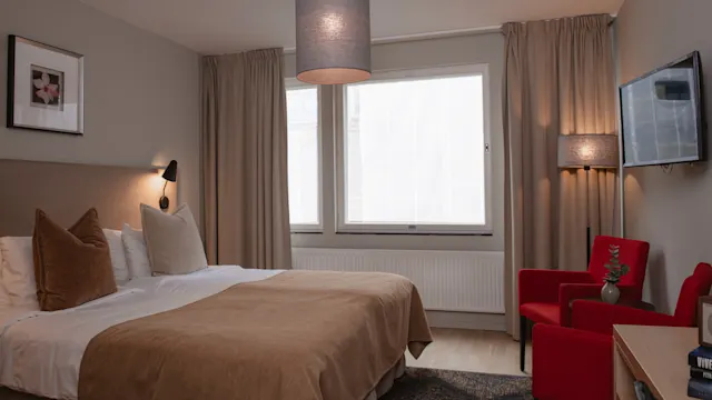 En noggrant uppbäddad säng med beige sängkläder och kuddar står mot en vägg i ett mysigt sovrum. Två stolar och ett bord finns vid sidan under en väggmonterad TV.