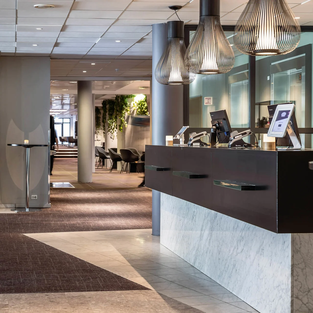 Reception placeret i en moderne hotellobby med computere på; tæt ved, en tændt pejs under et TV, og en gang der fører til siddeområder med grønt som accent.