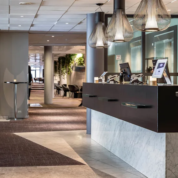 Resepsjonsdisk plassert i en moderne hotellobby med datamaskiner på; i nærheten, en tent peis under en TV, og en gang som fører til sittegrupper med grønne aksenter.