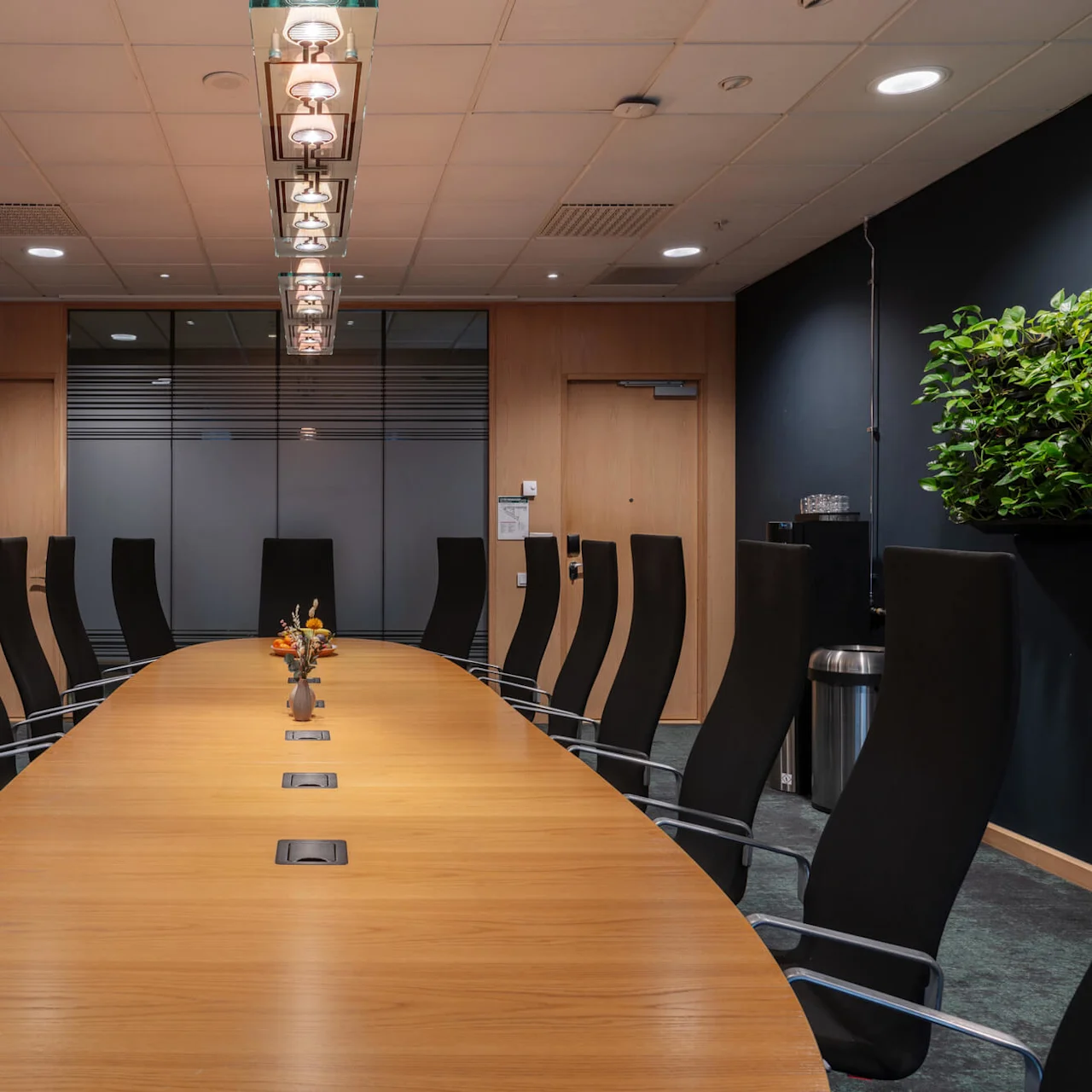 Ett långt träkonferensbord omgivet av höga svarta stolar har en vas med blommor, beläget i ett modernt mötesrum med mörka väggar och taklampor.