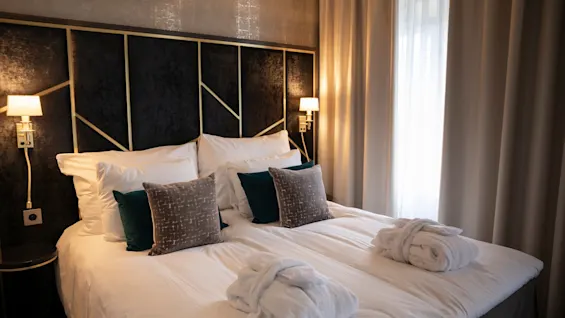 Et hyggeligt hotelværelse med en pænt redt seng, bløde puder og to hvide badekåber, der indbyder til afslapning og komfort.