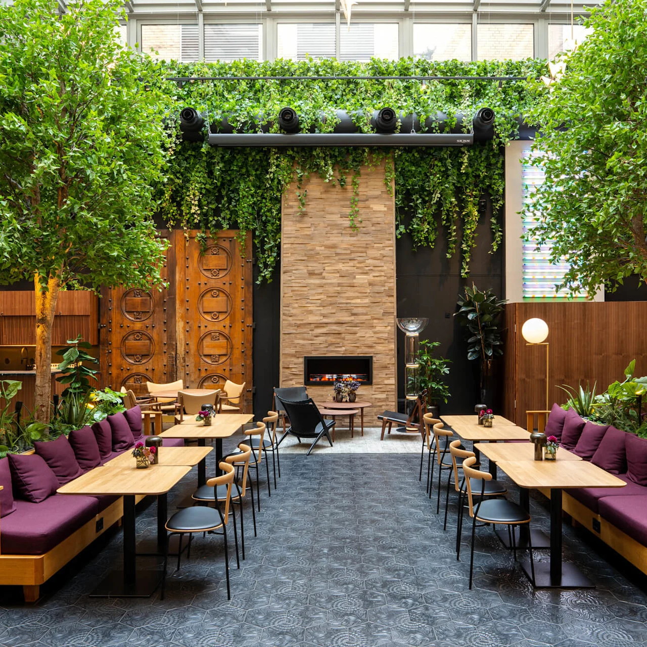 Seating area features wooden tables with chairs, surrounded by potted plants and trees under a glass ceiling. In the background, a stone wall and fireplace create a cozy ambiance.