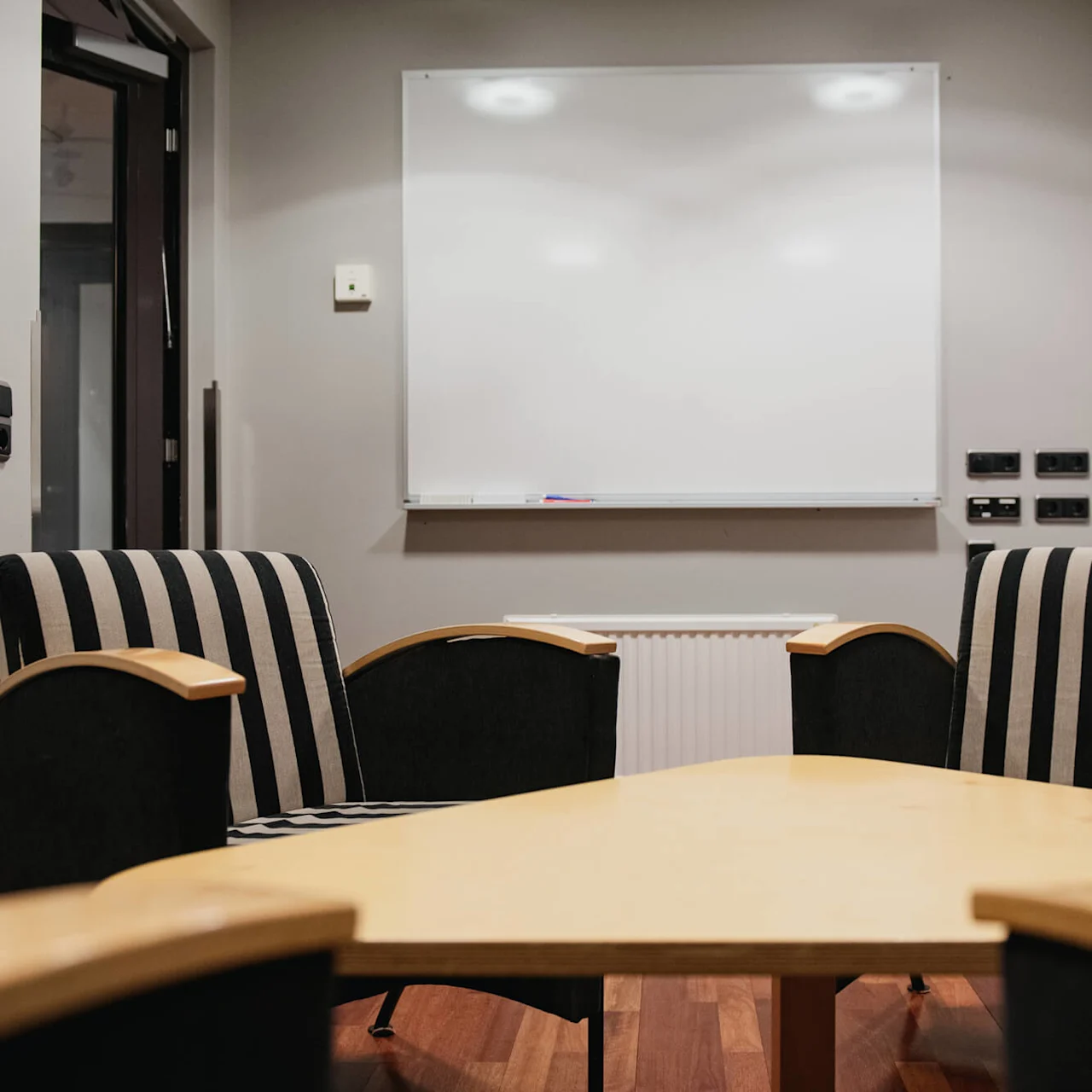 A wooden conference table faces striped chairs, set against a gray wall with a blank whiteboard, located in a meeting room.
