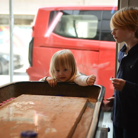 A young child leans over a table, reaching with curiosity, while an older child stands nearby holding an object. They are indoors near a window with a red vehicle outside.
