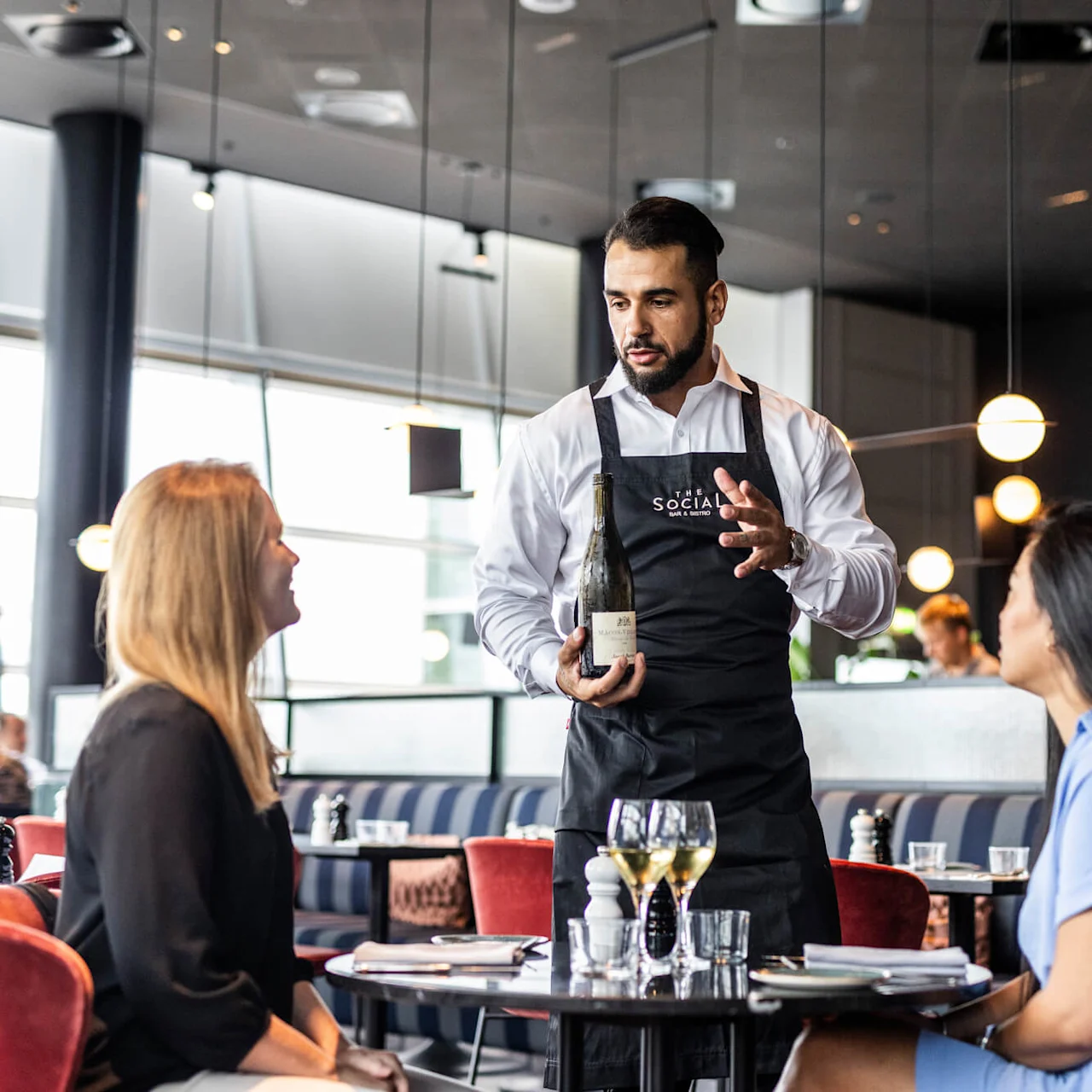En servitör i svart förkläde presenterar en vinflaska för två sittande kvinnor i en modern restaurang med runda lampor. Förklädestext: "THE SOCIAL".