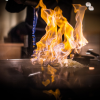 Flames engulf food being cooked on a flat grill, chef's hands visible, in a dimly lit restaurant setting.