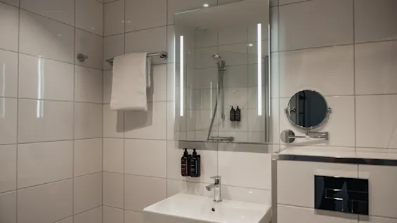 Ett rent, modernt badrum med vita kaklade väggar, ett handfat, en upplyst spegel och en handduk. En dusch reflekteras i spegeln.