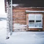 En rustik tømmerhytte delvist dækket af sne står isoleret i et snedækket landskab, med et snelandskab der strækker sig mod nåleskov under en bleg himmel.
