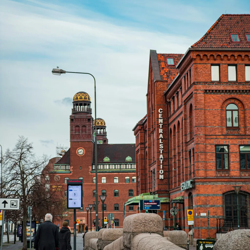 Folk går mot Sentralstasjonen i Malmö, en stor mursteinsbygning med et klokketårn under en blå himmel.