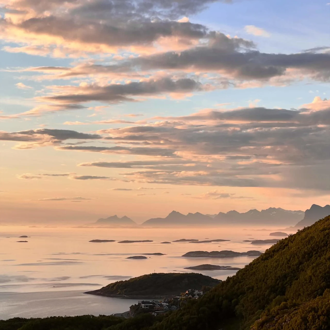 Sky illuminated by a soft sunset glow, clouds drifting gently above a serene coastal landscape with lush green hills and distant mountains, under a peaceful, evening sky.