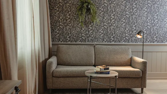A cozy living area with a sofa, books on a table, and a floor lamp, set against a patterned wall.