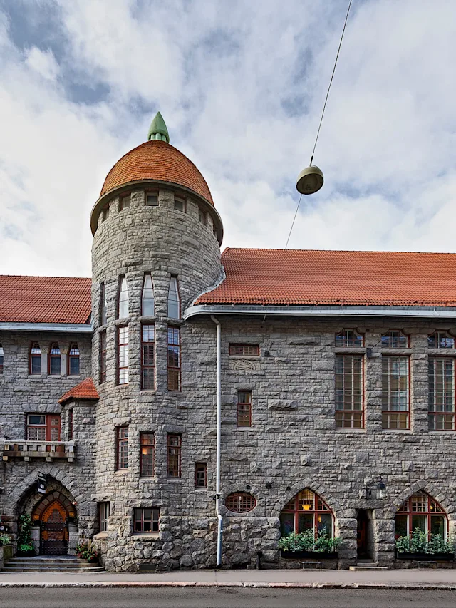 En historisk steinbygning har et sylindrisk tårn med rødt tegltak, plassert i en urban gatekontekst under en skyet himmel.