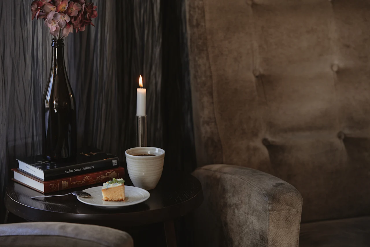 En mysig scen med ett tänt ljus, kaffe och kaka på ett bord bredvid en fåtölj. Böcker med titeln Malin Nord Barmark är staplade.