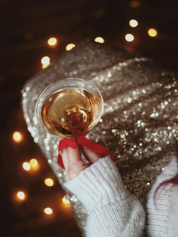 En hånd i en varm genser holder et festlig glass musserende drikke med et rødt bånd, blant festlige lys.
