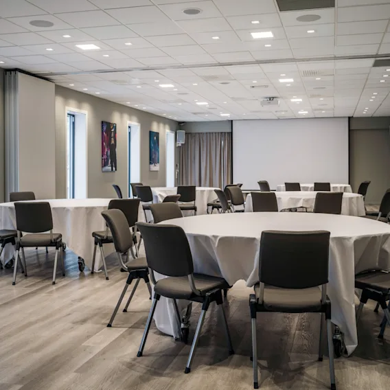 Et innbydende arrangementslokale med runde bord dekket i hvitt, klart for en samling eller presentasjon, komplett med en stor skjerm.
