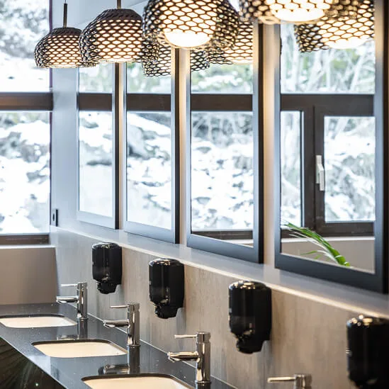 Sinks are lined up with mirrors above them, under decorative ceiling lights. Soap dispensers are mounted on the wall, and large windows reveal a snowy outdoor scene with plants inside.