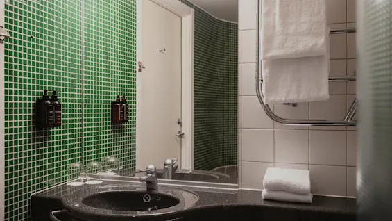 A well-lit bathroom at Home Hotel Uppsala featuring green mosaic tiles, a dark sink, and fresh white towels.