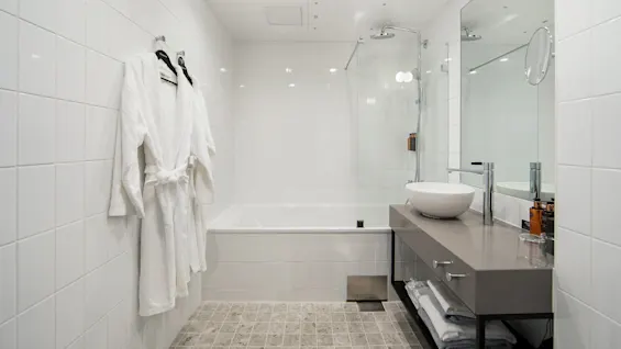 White bathrobe hangs on tiled wall, adjacent to bathtub with glass shower screen. Countertop holds a basin, faucet, and toiletries. Brightly lit, minimalist bathroom features folded towels below.