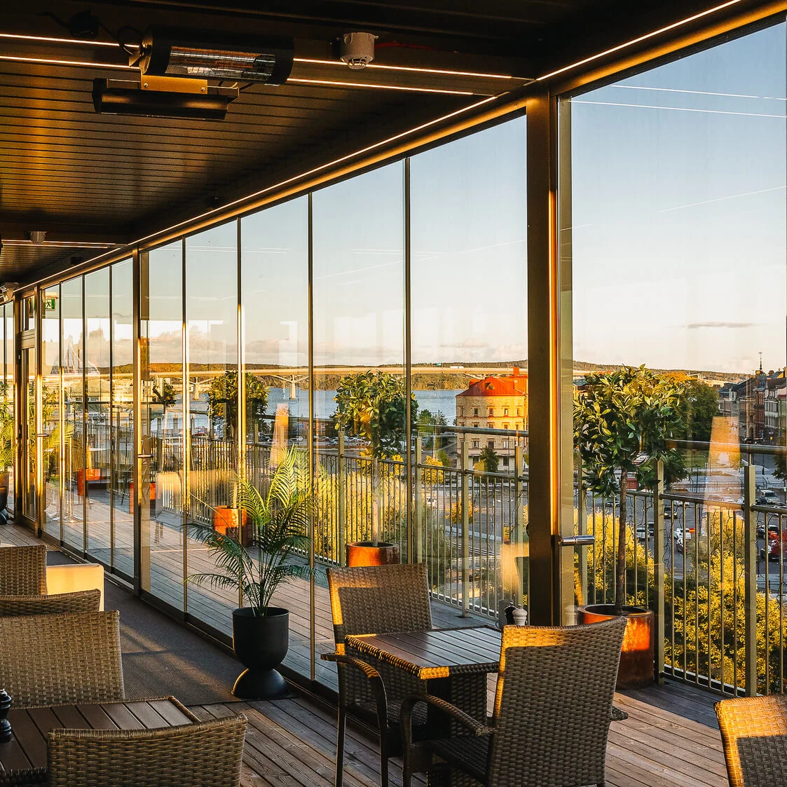 Rummelig terrasse med kurvestole og borde, med udsigt over et livligt bylandskab gennem glasvægge i solnedgangen. Lush planter og blød belysning skaber en hyggelig atmosfære.