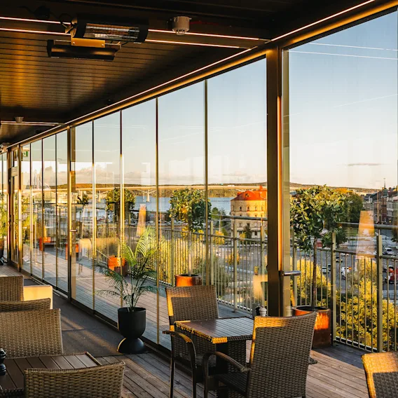 Spacious terrace with wicker chairs and tables, overlooking a vibrant cityscape through glass walls during sunset. Lush plants and soft lighting create a cozy atmosphere.