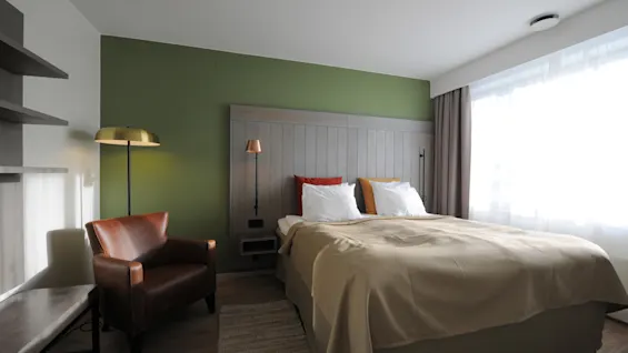 A neatly made bed with beige linens is set against a green accent wall. An armchair and floor lamp stand nearby. Sunlight filters through large curtains.