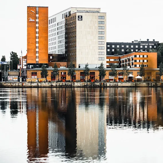 Höga byggnader speglas i en lugn vattenyta, med logotyper för "Diös", "Comfort Hotel" och "Scandic", mitt i ett stadslandskap med träd och en båt.