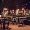 A restaurant features wooden tables and chairs arranged neatly, surrounded by soft lighting. Indoor plants adorn the space, creating a cozy ambiance with warm tones and modern decor.