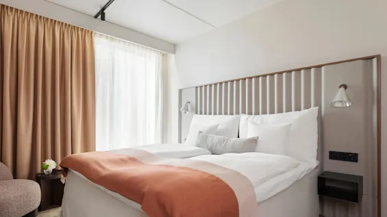 Säng täckt med vit sängkläder och en orange filt står i ett mjukt upplyst rum. Beige gardiner täcker stora fönster bredvid ett litet bord med en blomma.