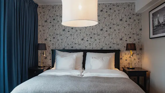 A welcoming hotel bedroom featuring a comfortable bed with white pillows, dark blue curtains, and elegant floral wallpaper.