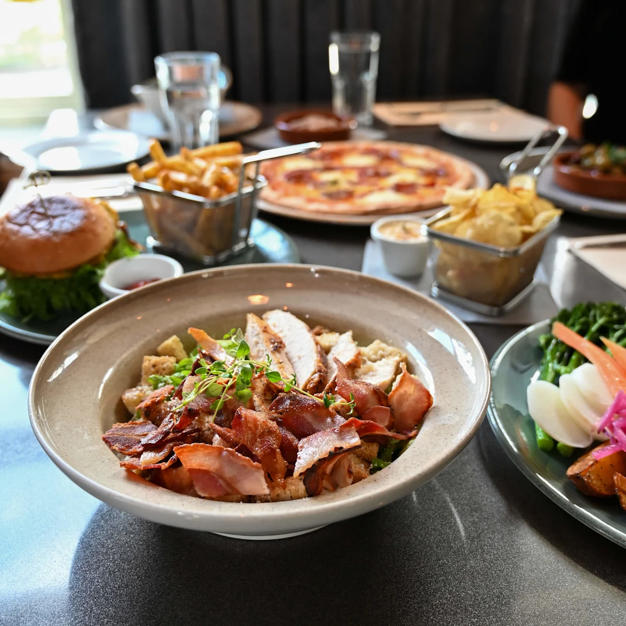 En skål med salat toppet med skiver af kylling og sprødt bacon, omgivet af forskellige retter inklusive en burger, pomfritter og pizza, på et mørkt restaurantbord.
