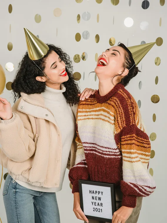 Two people in party hats laughing and celebrating New Year, holding a sign that says HAPPY NEW YEAR.