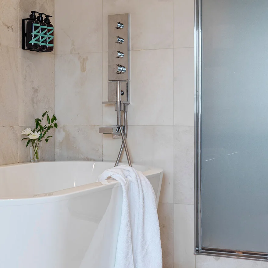 A white bathtub holds a towel, placed against a tiled bathroom wall. Above, shampoo and conditioner bottles are mounted. A small vase with flowers adds decor beside a modern shower fixture.