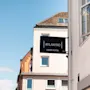 Et sort skilt med teksten HOME HOTEL er fastgjort til en hvid bygning, med et glimt af andre strukturer og en blå himmel.