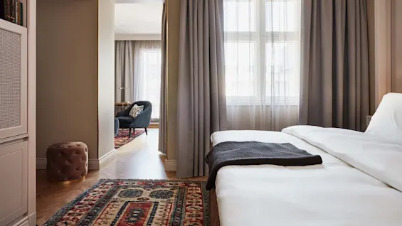 En pent oppredd seng med et grått teppe står i et koselig rom. Sollys strømmer gjennom gardiner og avslører et sitteområde med en stol og lampe utenfor.